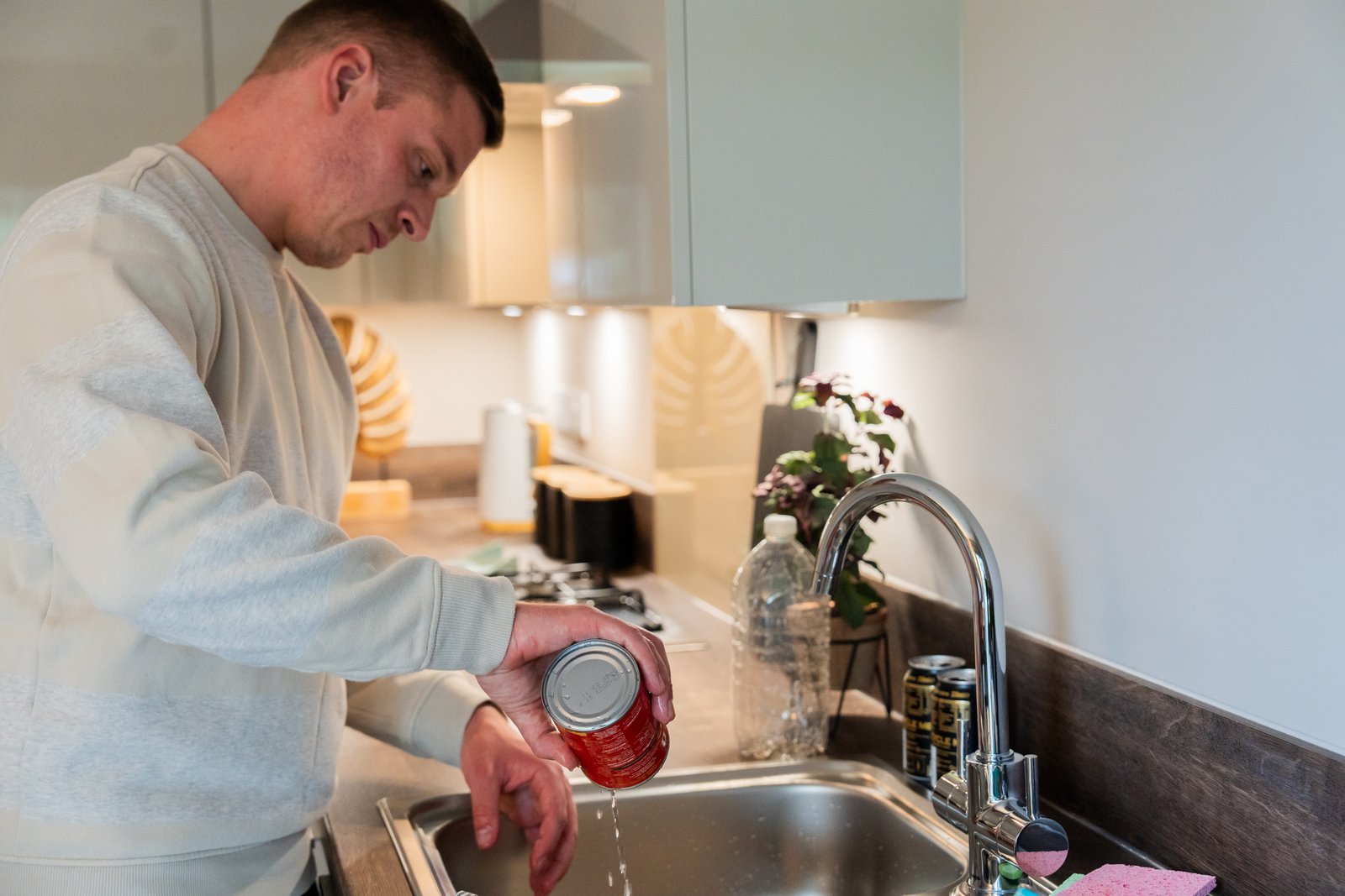 A man tips water from a food tin over the kitchen sink.